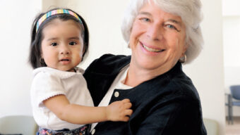 A smiling older woman with short gray hair holds a toddler girl with dark hair, wearing a headband and denim outfit. They are indoors, standing in front of a wall with the text "Marin Airporter & Hughes Family Fund." The scene captures an amazing moment of grace and warmth.