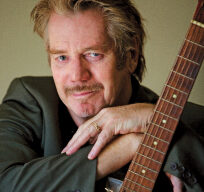 A man with light brown hair and a mustache, reminiscent of Dan Hicks, is posing with his arms crossed over a guitar. He is wearing a green blazer and smiling slightly at the camera. The background is plain and neutral-colored.