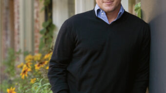 A man with short, styled hair stands outdoors, leaning against a wall. He is wearing a black sweater over a blue checkered shirt and blue jeans. Plants with yellow flowers are in the background along a walkway. He is smiling slightly and looking at the camera, resembling celebrity chef Tyler Florence.