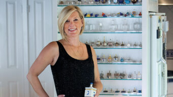 A woman with short blonde hair, wearing a sleeveless black top, stands in front of a display shelf filled with various small decorative items, including dreidels. She holds a small item in her left hand and smiles at the camera, embodying the vibrant energy of Spin City.