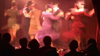 A lively tango performance on stage with couples dressed in colorful, elegant costumes dancing energetically. It truly takes two to tango, and the dancers' movements create a blur effect. The audience is seated, silhouetted in the foreground, watching the performance under dramatic lighting.