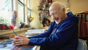 A smiling elderly man in a dark blue hoodie sits at a table doing arts and crafts, pouring his heart into each piece. Behind him, a young woman focuses on her own project. They are in a brightly lit room with colorful decorations and a large window.