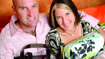 A man and a woman sit close together, smiling at the camera. They are surrounded by various patterned Fleurville bags. The man is wearing a light pink shirt, and the woman is in a black and white patterned top. The background features a warm, orange-toned design that complements their fashion-forward look.