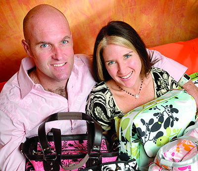 A man and a woman sit close together, smiling at the camera. They are surrounded by various patterned Fleurville bags. The man is wearing a light pink shirt, and the woman is in a black and white patterned top. The background features a warm, orange-toned design that complements their fashion-forward look.
