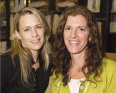 Two women smiling at the camera. The woman on the left, from an eco-friendly mom network, has blonde hair and is wearing a dark top, while the woman on the right, who supports environmental sustainability, has curly dark hair and is wearing a green jacket over a white shirt. They are standing in front of a shelf with items on it.