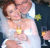 A bride and groom are joyfully embracing, each holding a glass of champagne. The bride is wearing a white wedding dress and veil, while the groom is in a dark suit with a boutonniere. Both are smiling and appear to be celebrating their Encore Celebrations wedding.