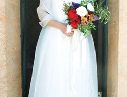 A woman in a sleeveless white gown and light shawl stands holding a colorful bouquet of flowers. She is standing in a doorway, smiling at the camera, embodying the essence of timeless style.