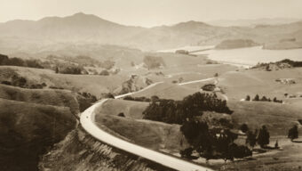 A winding road snakes through a vast, hilly landscape under a hazy sky. The terrain is dotted with trees and patches of vegetation, with mountains visible in the distance. This serene and expansive scenery not only showcases the natural beauty of the countryside but also hints at its rich road development history.