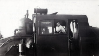 Black and white photo of a man named Provines sitting inside the cab of steam locomotive number "8," smiling and looking out of the side window. The locomotive features visible mechanical parts and a chimney.