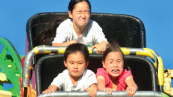 Three children with expressive faces ride a roller coaster in County Fear. The child in front, dressed in a red shirt, grips the bar tightly, showing a mix of excitement and horror. The other two children behind also exhibit similar intense expressions, highlighting the thrill of the ride.