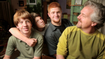 A group of four people, composed of three young men and one older man, smile and embrace each other. They are indoors in a warmly lit room with green walls and framed pictures in the background. The atmosphere appears friendly and familial, with the "Green Dad" proudly at the center.