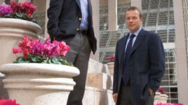 Two men in business attire stand on stone steps adorned with flower pots filled with vibrant pink flowers. The modern building behind them, a symbol of equity, features tall columns and large glass windows. One man stands on a higher step, and the other is slightly below him, demonstrating access for all levels.