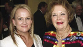 Two women are standing together and smiling at a formal event in Marin County. The woman on the left has light hair and is wearing a white outfit, while the woman on the right has curly hair and is wearing a colorful, striped jacket. People are mingling in the background, likely anticipating a performance by the Marin Symphony.