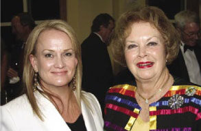 Two women are standing together and smiling at a formal event in Marin County. The woman on the left has light hair and is wearing a white outfit, while the woman on the right has curly hair and is wearing a colorful, striped jacket. People are mingling in the background, likely anticipating a performance by the Marin Symphony.