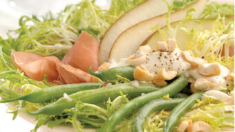 A fresh Haricot Vert Salad featuring pear slices, thinly sliced Serrano ham, green beans, and frisée lettuce, garnished with chopped nuts and drizzled with a creamy dressing. A wine glass and a neutral tablecloth are blurred in the background.