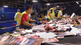 Workers in safety vests sort through various papers and cardboard on a moving conveyor belt at the Marin Recycling Center. The background shows machinery and more recyclable materials.