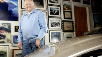An older man stands in a room filled with framed pictures and memorabilia of speedboats and racing, including tributes to Howard Arneson. He is wearing a light blue sweater and jeans. Part of a polished car is visible in the foreground. The man is smiling and has one hand in his pocket.