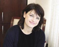A person with shoulder-length dark hair and a friendly smile is sitting indoors. They exude beauty, wearing a dark-colored top, positioned in front of a wooden cabinet and light-colored curtains.