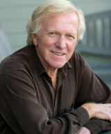 An older man with light-colored hair smiles while leaning on a railing. He is wearing a dark brown shirt and has his arms crossed. The background is blurred with fresh light-colored siding and some outdoor furniture visible.