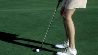 A person standing on a golf course is about to putt a golf ball into the hole. The golfer, looking fresh after a quick makeover, is wearing light-colored shorts and white golf shoes with gray and green detailing. Only the lower half of the body is visible, with focus on the golf ball and putter.