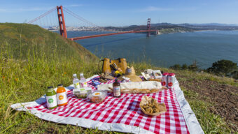 A red and white checkered picnic blanket is spread on a grassy hill, overlooking the Golden Gate Bridge and San Francisco Bay. The blanket has various picnic picks, food items, drinks, and a wicker basket. The sky is clear and blue, offering a scenic view of the city and bridge.
