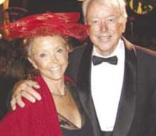 An older couple stands together, smiling. The woman wears a large red hat, a red shawl, and a black dress. The man is dressed in a black tuxedo with a bow tie. They are posing in a warmly lit setting, possibly at a formal event, embodying the spirit of La Dolce Vita.