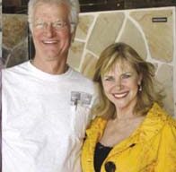 A smiling man and woman stand side by side at an Open House event. The man wears glasses and a white shirt, while the woman sports a bright yellow jacket adorned with a small shamrock pin. They are indoors with a tile-patterned wall in the background, captured warmly from the waist up.