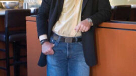 A man is standing and leaning against an orange-colored counter in a modern kitchen, embodying a chic lifestyle. He is wearing a light yellow shirt, blue jeans, and a black blazer, smiling as he holds a cigarette. The background includes bar stools, kitchen equipment, and hanging lights.