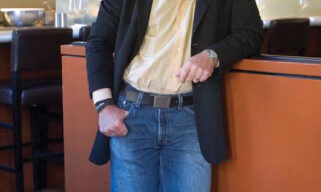 A man is standing and leaning against an orange-colored counter in a modern kitchen, embodying a chic lifestyle. He is wearing a light yellow shirt, blue jeans, and a black blazer, smiling as he holds a cigarette. The background includes bar stools, kitchen equipment, and hanging lights.