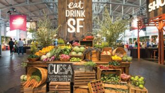 A vibrant market display features a variety of fresh fruits and vegetables arranged on wooden crates and tables. A large sign reads "eat DRINK SF" above the produce, celebrating our love for food festivals, with another sign for CUESA promoting a healthy food system. People are visible in the background.