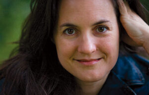 A close-up shot of a person with long dark hair, wearing a dark jacket, smiling gently at the camera. Their head is slightly tilted to one side with one hand resting against their cheek. The background is soft-focused greenery, reminiscent of where advocacy director Deb Hubsmith might have enjoyed nature.