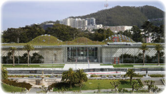 The image shows a modern building with a green rooftop, set against a backdrop of trees and hills. The facade is mostly glass, allowing views into the interior. In front of the building, there are well-maintained gardens and pathways where local happenings often take place.