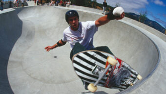 A skateboarder wearing a helmet and protective gear rides up the curved wall of a large, concrete skatepark bowl in Marin. The sky is blue with some clouds, and a few people are gathered in the background watching.