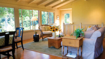 A cozy living room with a vaulted ceiling, wooden beams, and large windows. It features a sofa, wicker chair, and coffee table with sunflowers. A green rug covers part of the hardwood floor. Carefully staged for sale, the small dining area with a table and chairs sits invitingly in the corner.