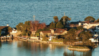 A coastal residential area in the foreground features a collection of houses surrounded by trees and a calm body of water. The houses, built close to the water's edge, offer a belvedere for residents, with several birds visible on the water's surface and the open sea in the background.