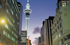 A bustling city street at dusk, lined with tall buildings on both sides, exudes Kiwi Chic. The prominent Sky Tower stands illuminated in the distance. Blurred lights from moving vehicles create dynamic streaks, indicating traffic. The sky is transitioning from light to dark.
