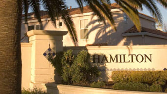 A tall palm tree and lush greenery in the foreground partially shadow a beige sign with "Hamilton" written on it. The sign appears to be part of a wall encircling a residential area, reminiscent of a Broadway set, with a two-story house visible in the background.