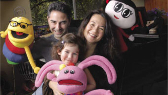 A family of three, consisting of two adults and one child, smiles at the camera. They hold colorful plush toys, including a yellow insect, a pink rabbit, and a character with black hair and a red dress like Dora. A piano is visible in the background.