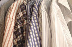 A row of clean collared shirts neatly hung on hangers. The shirts feature various patterns including stripes and checks in colors such as red, blue, and gray. The hangers are placed on a wooden rod. The background is softly blurred with a hint of green, eco-friendly ambiance.