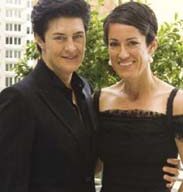Two women in formal attire stand close together, smiling warmly at the camera. The woman on the left has short dark hair and wears a black suit, while the woman on the right, also with short dark hair, is dressed in an off-the-shoulder black dress. They are outdoors with buildings in the background, representing Sunny Hills Services with grace and professionalism.
