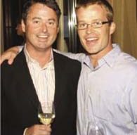 Two men smiling and posing together indoors. The man on the left, wearing a dark jacket, holds a glass of white wine. The man on the right, wearing a light blue shirt and glasses, holds a glass of red wine in one hand and has his arm around the other man's shoulder at the Bay Institute event.