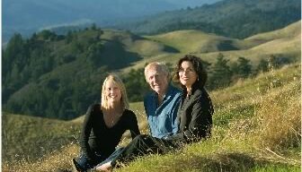 Three people are sitting on a grassy hillside with rolling hills and forests in the background. They are smiling and enjoying the scenic view of Mt. Tam, bathed in natural light.