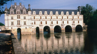 A grand, historic chateau in the Loire Valley with numerous windows and arched supports sits over a calm river on a sunny day. Trees line the water's edge, and the reflection of the building is visible in the water below, making it a picturesque spot for tourism in France.