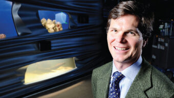 A smiling man with a mission, dressed in a green blazer and blue tie, stands confidently in front of a display of various objects. The background features a dark, wavy structure with visible items like rocks and a white illuminated section.