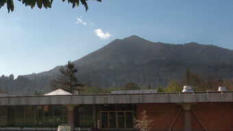 A modern, low-rise school building with large windows sits on a well-maintained grassy area. Behind it, a tree's leaves frame the image from the top. In the background, a tall, forested mountain rises under a clear blue sky, symbolizing new beginnings and second chances.