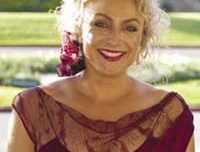 A woman with curly blond hair, wearing a red dress with lace details, smiles while standing outdoors on a sunny day. She has red lipstick and a flower in her hair, framed as if ready for "lights, camera, action" against the blurred backdrop of greenery.