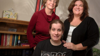 Three women are indoors posing for a photo. Two women stand behind a seated younger woman, all looking good. The woman on the left wears a red sweater, the woman on the right, possibly one of the lawyers, wears a black cardigan, and the seated woman sports a black sports-themed shirt. A bookshelf is in the background.