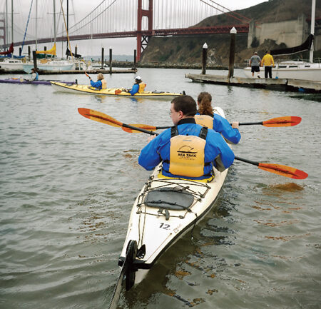 A group of kayakers in blue jackets and yellow life vests paddle through a marina with docks and boats, all along for the glide. The iconic Golden Gate Bridge is visible in the background under a cloudy sky.