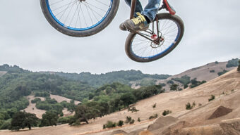 A person wearing a helmet is performing an aerial trick on a bicycle against a backdrop of hills and cloudy sky at Stafford Lake Bike Park. The rider is mid-jump and the bicycle is tilted, capturing the action of a dirt trail park.