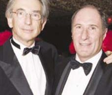 Two men dressed in formal black tuxedos with black bow ties are posing together at a San Francisco Symphony event. Both have light-colored hair; one is wearing glasses. They are smiling and standing close to each other, with their arms around each other's shoulders.